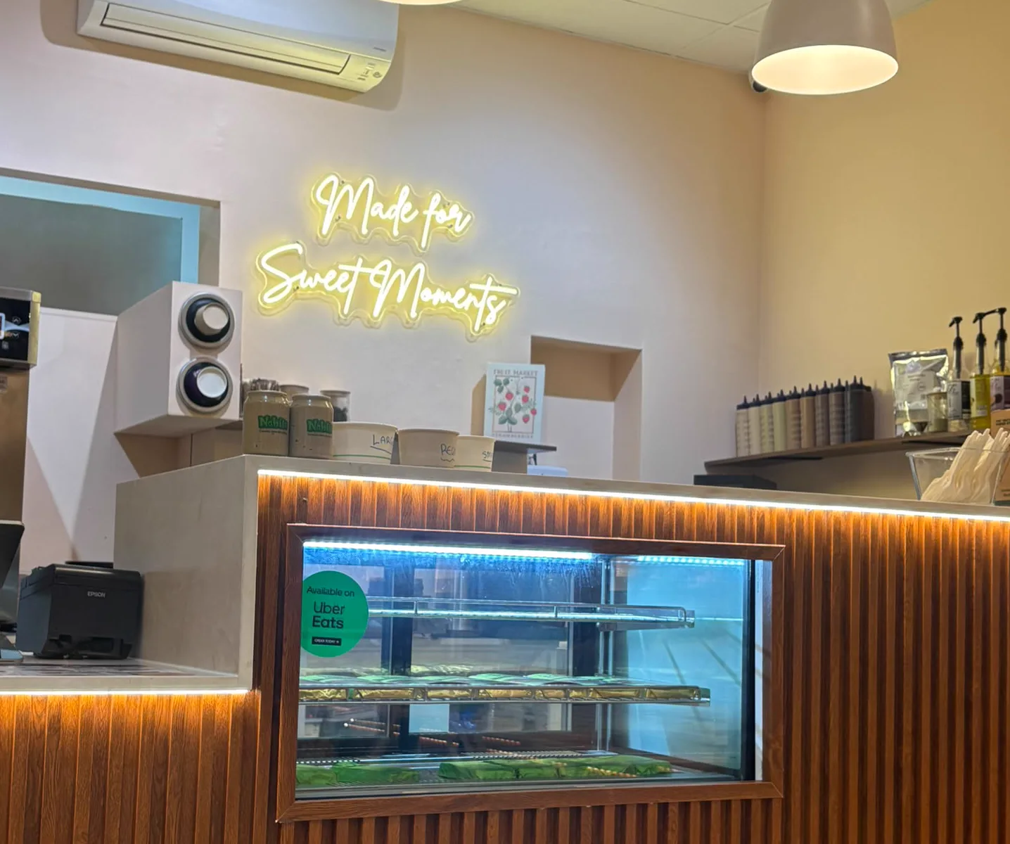 A neon sign reading “Made for Sweet Moments” glowing on the wall above the counter at Nabil's Açaí Station in Mount Lawley, with topping jars on the shelf and the lit display cabinet below.